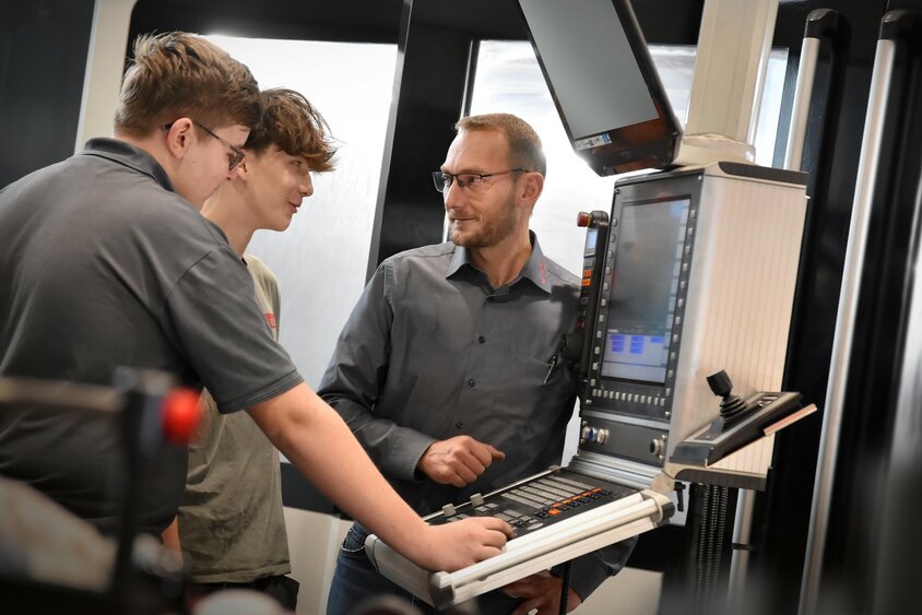 Ein Lehrlingsausbilder erklärt zwei Lehrlingen im Bereich Metalltechnik, Modul H8 Zerspanungstechnik, die Steuerung eines Bearbeitungszentrums. Die Lehre in Steyr bei weba kombiniert praktische Erfahrung mit theoretischen Know-how in der Zerspanungstechnik.