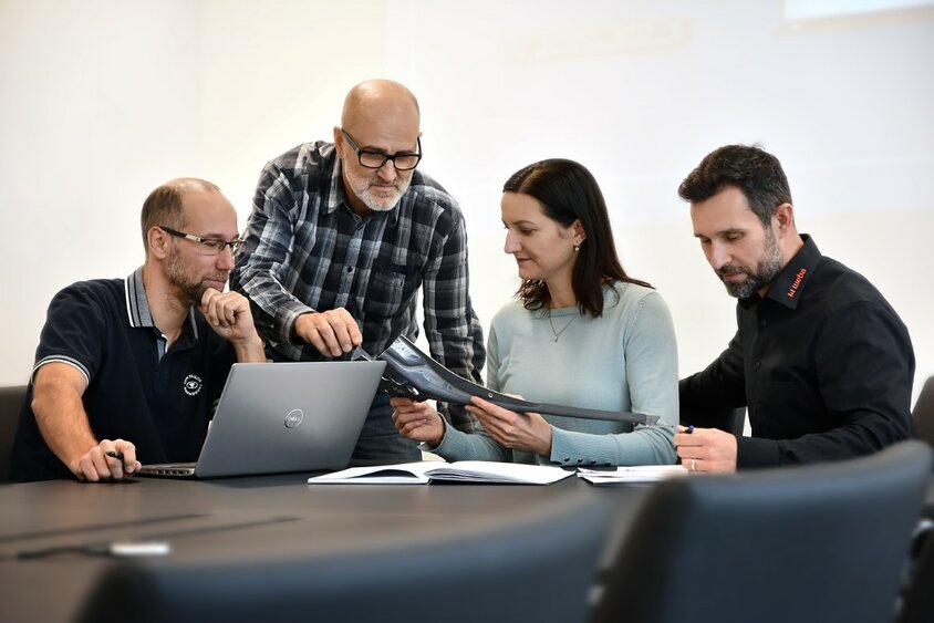 Three male and one female employee of weba Werkzeugbau are sitting in a meeting discussing a steel component. A laptop and documents are on the table.