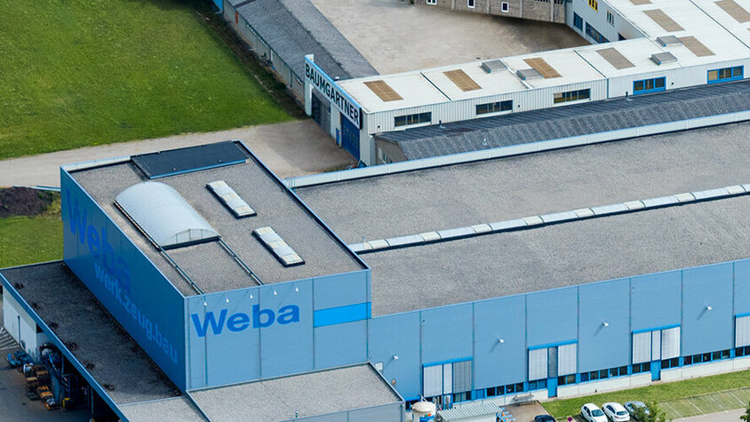 Die Luftaufnahme zeigt das beeindruckende Headquarters und Entwicklungszentrum von weba in Dietach. Die Produktionsstätte erstreckt sich über das Gelände, während im Vordergrund die Pressenhalle mit der deutlich sichtbaren Beschriftung "WEBA" zu sehen ist. Die Architektur wirkt modern und funktional, während die Umgebung von Grünflächen und Straßen umgeben ist. Die Szene vermittelt einen Eindruck von Größe und Professionalität, während das "WEBA"-Logo die Identität des Unternehmens betont.