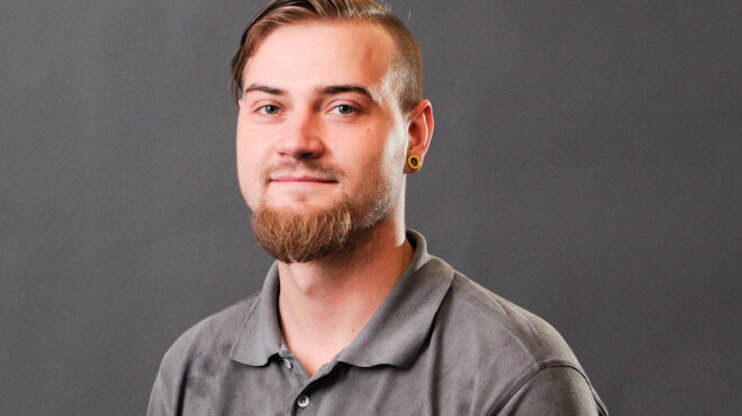 [Translate to English:] Porträtfoto von Bronislav Škoda, einem jungen Mann mit kurzem, seitlich rasiertem Haar und einem gepflegten Vollbart. Er trägt ein graues Poloshirt mit dem Logo von weba und schaut freundlich in die Kamera. Seine Ohren sind mit gelben Plugs geschmückt, und im Hintergrund ist eine neutrale graue Studioleinwand zu sehen.