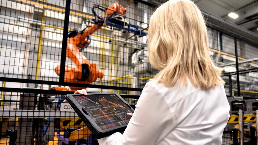 Eine Frau steht in einer industriellen Umgebung bei weba Werkzeugbau vor einem großen Robotergreifer einer Warmumformlinie. In ihren Händen hält sie ein Tablet, auf dem sie Daten überwacht oder Steuerbefehle für den Roboter eingibt. Der Fokus liegt auf der Interaktion zwischen moderner Technologie und menschlichem Eingreifen.