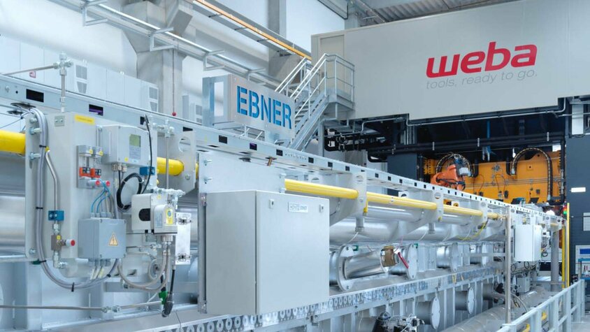 Foto einer Warmumformanlage von weba Werkzeugbau, auf dem ein 22 Meter langer Rollenherdofen und eine 1.600 Tonnen Umformpresse zu sehen sind. Die Anlage zeigt die industrielle Fertigungstechnik der Warmumformung in Aktion, wobei der Ofen für die Erwärmung von Metallplatten vor der Umformung verwendet wird und die Presse das erwärmte Metall in die gewünschte Form bringt.