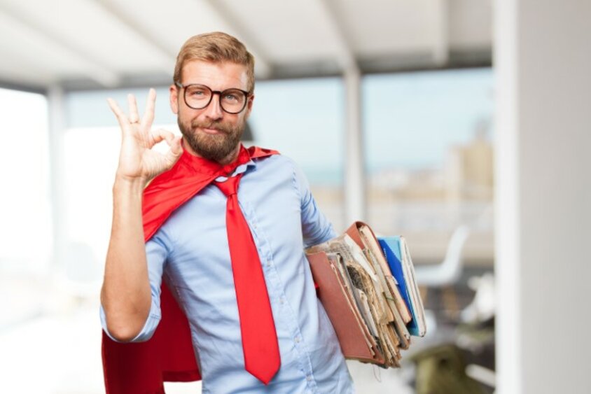 A man in a suit with a red cape and red tie holds files and makes an OK sign to signal approval and competence.