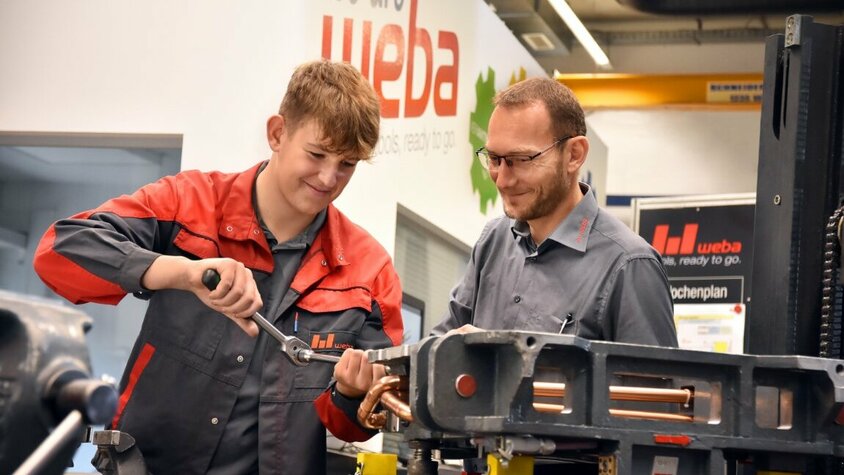 Ein Lehrling in der Werkzeugbautechnik arbeitet an einer Miniatur eines Umformwerkzeugs, während der Ausbilder aufmerksam zuschaut. weba Werkzeugbau bietet Lehre in Steyr in der Metalltechnik an.